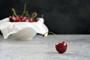 Tek bir kiraz gri mermer yüzey üzerinde yatıyor ve odaklanmıştır, arka planda bir kase birçok kiraz vardır 