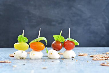 Farklı renklerde küçük domates bir kürçat ile mozzarella topu üzerinde sıkışmış, üstüne bir fesleğen yaprağı döşenmiştir - Gri mermer yüzeyüzerinde vitamin çok küçük taze çerez