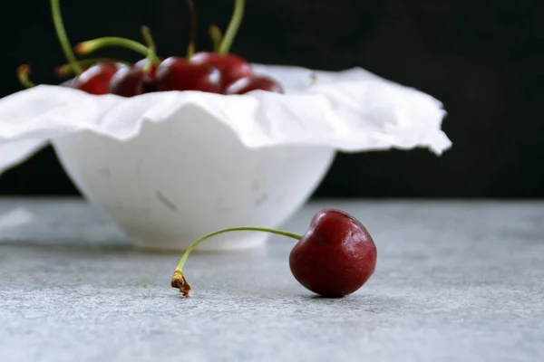 A single cherry lies on a gray marble surface and is focussed, in the background are many cherries in a bowl