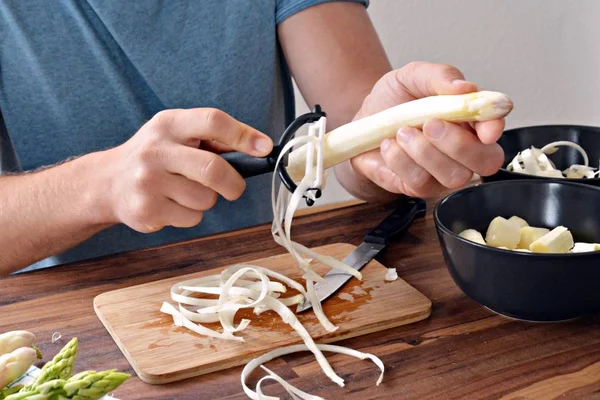 A person peels fresh white asparagus with a small peeler. The board was photographed with the hands of the person