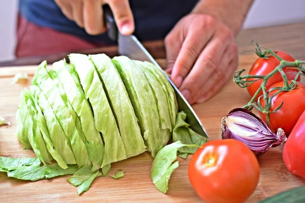 A person cuts a half head of lettuce on a wooden board for a fresh crisp healthy salad - close-up on the hands of the person - concept for healthy nutrition with fresh organic vegetables