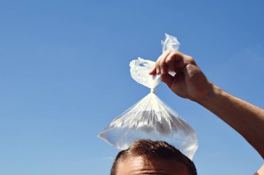 Yirmili yaşlarda bir adam bulutlar olmadan berrak bir mavi gökyüzü önünde soğuk su dolu bir plastik torba ile başını soğutur - Avrupa'da sıcak yaz serinletici soğutma 