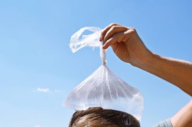 Yirmili yaşlarda bir adam bulutlar olmadan berrak bir mavi gökyüzü önünde soğuk su dolu bir plastik torba ile başını soğutur - Avrupa'da sıcak yaz serinletici soğutma 