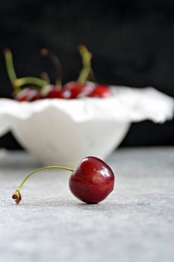 Tek bir kiraz gri mermer yüzey üzerinde yatıyor ve odaklanmıştır, arka planda bir kase birçok kiraz vardır 