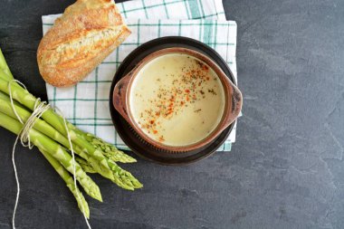 Kuşkonmaz kremaçorbası dolu bir porselen tencere koyu mermer bir yüzey üzerinde duruyor, yanında bir çay havlusu ve bir rulo ile 