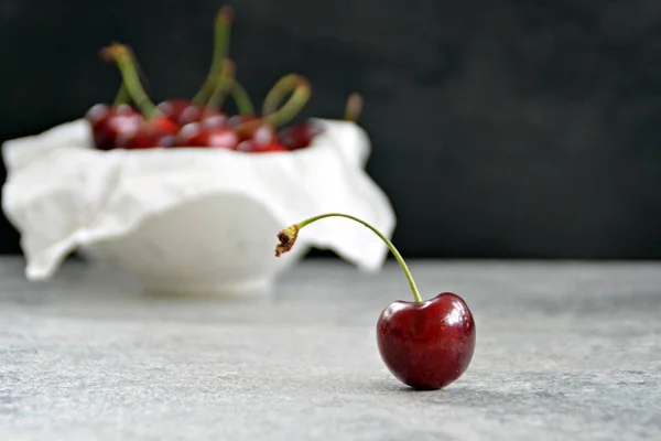 A single cherry lies on a gray marble surface and is focussed, in the background are many cherries in a bowl