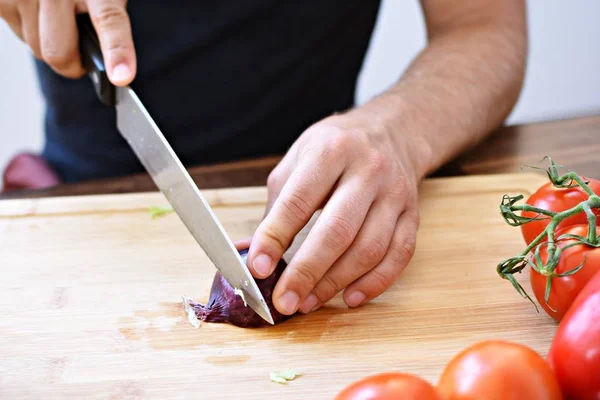 A person cuts a red onion on a wooden board for a fresh crunchy healthy salad - close-up on the hands of the person - concept for healthy nutrition with fresh organic vegetables