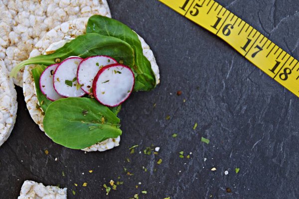 On a dark marble surface are three round rice waffles, one with lettuce and radishes and a yellow tape measure with space for text or other elements - concept for healthy nutrition in the gym