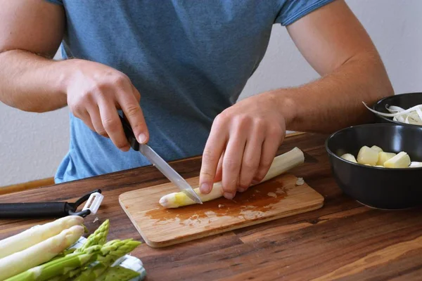 A person peels fresh white asparagus with a small peeler. The board was photographed with the hands of the person