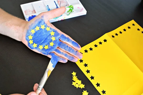 A person is holding up his hand in front of a cloudy sky , the hand is painted with the colors of the european union with yellow paper stars - concept to celebrate the eu and the freedom of vote