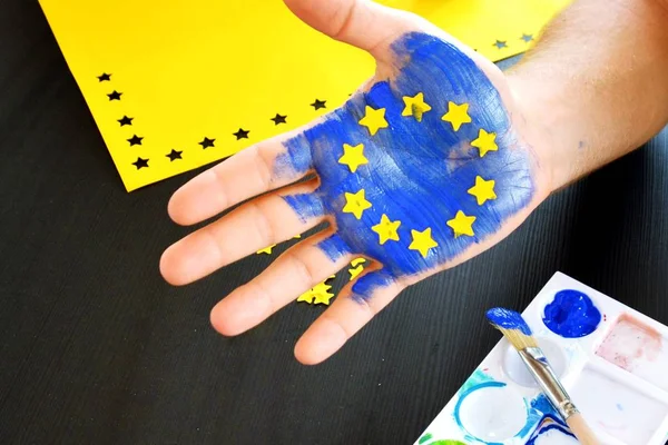 A person is holding up his hand in front of a cloudy sky , the hand is painted with the colors of the european union with yellow paper stars - concept to celebrate the eu and the freedom of vote