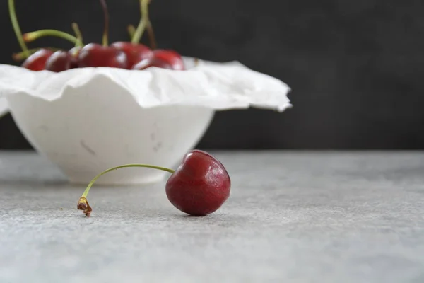 A single cherry lies on a gray marble surface and is focussed, in the background are many cherries in a bowl