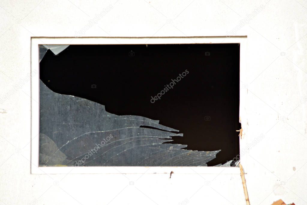 A destroyed glass pane, photographed from outside, a very dark room behind it - a white wall with a broken window in it