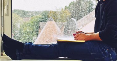 Bir adam pencerenin kenarında sıcak bir bardak içecekle oturuyor, pencere camındaki yağmur damlalarında, kitap okuyor ve sonbaharda yağmurun ve sessizliğin tadını çıkarıyor.