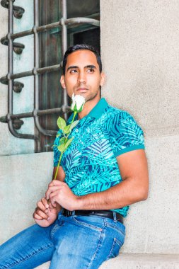 Genç Latin Amerikalı beyaz holding adam sen, yeşil desenli Polo gömlek giymiş sizi eksik gül, New York'ta sokakta pencere ile eski duvara oturmuş, uzağa arıyorsunuz, düşünme