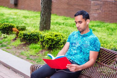 Genç Latin Amerikalı adam, yeşil desenli Polo gömlek giymiş siyah pantolon, beyaz kulaklık, New York'ta, seyir, okuma kırmızı kitap, müzik dinleme sokak Park bankta oturmuş kablolu.