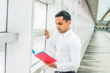 Genç Latin Amerikalı adam beyaz gömlek giymiş kırmızı kitap, NewYork'ta, üzerinde geçit üzerinde cam duvarın içinde bina, aşağı bakıyor, okuma, düşünme office tarafından ayakta