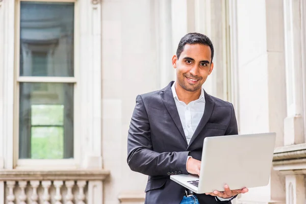 Young Handsome Hispanic American college student studying in New York, wearing black blazer, white shirt, sitting inside old office building on campus, working on laptop computer, looking up, smiling