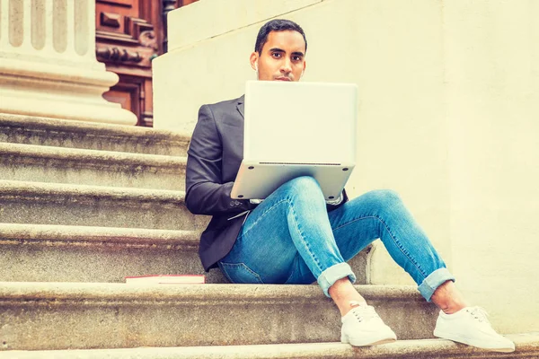 Young Hispanic American college student studying in New York, wearing black blazer, blue jeans, sneakers, earphone, sitting on stairs of office building, listening music, working on laptop computer