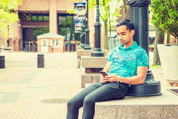 Young Hispanic American College Student, wearing green patterned Polo shirt, black pants, sitting against light pole on campus in New York, looking down, reading, texting on cell phone