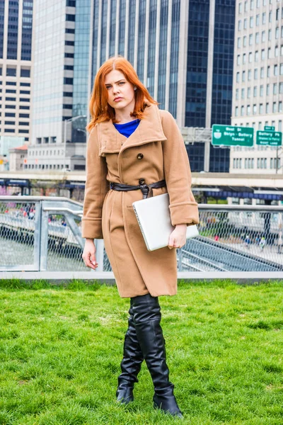 若い実業家旅行 冬のニューヨークの仕事長い茶色のウールコート 長い黒革のブーツを身に着けている 高層ビルとビジネス街の緑の芝生の上に立って — ストック写真