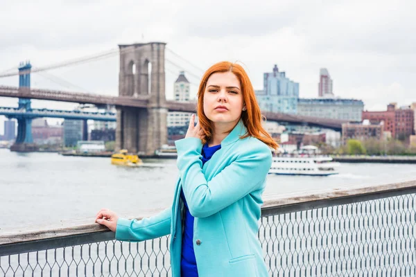 Young American Woman traveling in New York, wearing cadet blue woolen coat, blue shirt, standing by East River, narrowing eyes, hand touching hair. Manhattan, Brooklyn bridges, boats on background