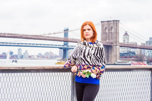 Young American Woman missing you, waiting for you, holding white rose, wrapping around body with patterned scarf in cold weather, frowned, thinking. Manhattan, Brooklyn bridges on background.