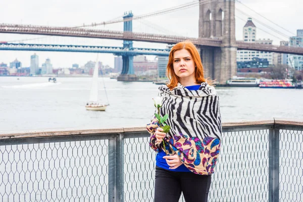 Young American Woman missing you, waiting for you, holding white rose, wrapping around body with patterned scarf in cold weather, frowned, thinking. Manhattan, Brooklyn bridges on background.