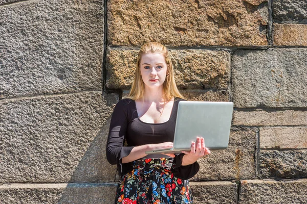 Young American teenage college student studying in New York, wearing black long sleeve low cut t shirt, holding laptop computer, standing against rock wall on campus under sun, looking, thinking
