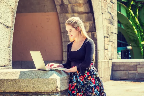 Young American teenage college student studying in New York, wearing black long sleeve t shirt, patterned skit, standing by rock gate on campus under sun, looking down, working on laptop computer