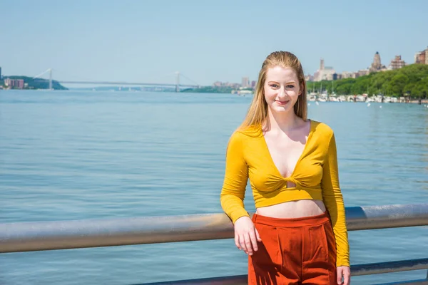 American teenage girl traveling in New York in spring, wearing yellow long sleeve deep v neck fit crop top, red orange pants, standing by Hudson River, smiling. Bridge, harbor in far background