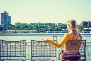 Boş sandalye sizin için. Yalnız kız bekleyen dostluk. Sarı uzun kollu giyiyor uygun ürün T gömlek, New York'ta Hudson Nehri tarafından sandalyeye oturmuş, New Jersey karşı karşıya uzun sarı saçlı Genç kadın.