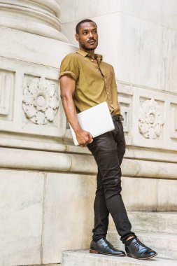 New York 'ta sakallı, yeşil kısa kollu gömlekli, siyah pantolonlu, deri ayakkabılı, dizüstü bilgisayar taşıyan, beyaz mermer duvarda duran Afrikalı genç bir üniversite öğrencisi.