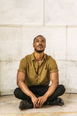 Sakallı, genç Afro-Amerikan Yeşil kısa kollu gömlek, siyah pantolon, deri ayakkabı giyen adam. bacak bacak, kampüs New York'ta duvara başına mermer yere oturmuş, baş yetiştirme, ararken