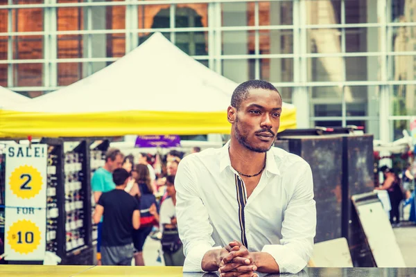 Summer Street Fair and Flea Market in New York. Young African American Man shopping, traveling in New York, wearing white shirt, standing on street in Midtown of Manhattan, thinking, lost in thought