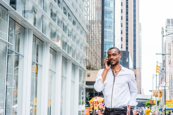 Young African American Man with beard traveling in New York, wearing white shirt, walking on street in Midtown of Manhattan, talking on cell phone. Modern high buildings, busy street on background