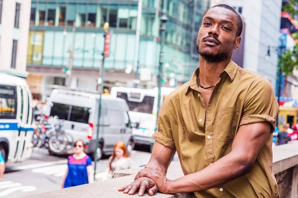 Lonely Man in Big City. Young African American Man wearing green short sleeve shirt, standing by half wall fence, facing busy street in Midtown of Manhattan, New York, sad, thinking, lost in thought