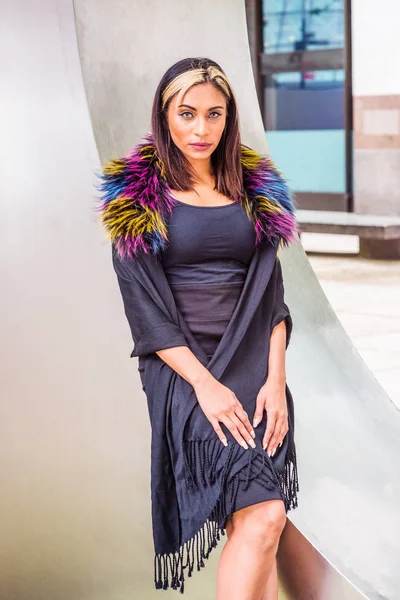 Portrait of Young American Woman in New York, with black hair, dyed front top little blonde, wearing black fit dress, wrapping shawl from shoulder down, colored faux fur scarf, standing by silver wall