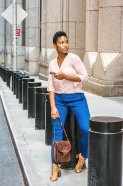 Genç Afro-Amerikan New York'ta kısa saç ile seyahat, pembe V boyun gömlek, mavi pantolon giyen kadın, kahverengi Peep yüksek topuklar, deri çanta, metal üzerinde oturan tutan sokakta, textIn ayağı.