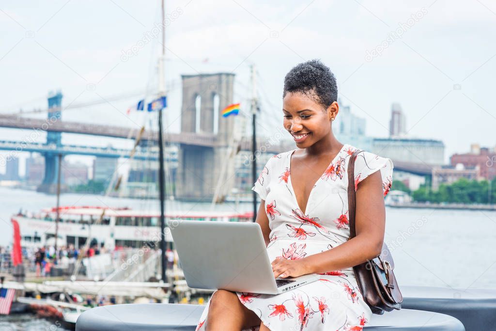 Young African American Woman traveling in New York, with short afro hair, wearing dress, carrying bag, sitting by river, working on laptop computer. Brooklyn, Manhattan bridges, boat on background