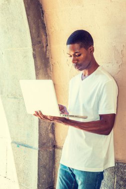 Genç Afro-Amerikan erkek üniversite öğrenci kampus kapısı tarafından yaz aylarında ayakta beyaz T gömlek giymiş eğitim New York City, kısa saçlı aşağı bakıyor, okuma, dizüstü bilgisayar üzerinde çalışan.