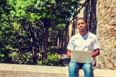 Dizüstü bilgisayar üzerinde çalışan genç Afro-Amerikan erkek üniversite öğrenci kampüse güneş altında sokakta tarafından yaz aylarında oturan beyaz T gömlek giymiş eğitim New York City, kısa saçlı, düşünme