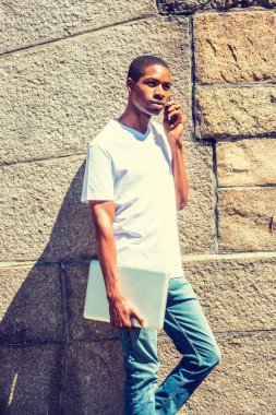 Genç Afro-Amerikan üniversite öğrenci dizüstü bilgisayar, yaz aylarında güneş altında kampüste kaya duvarı tarafından ayakta tutan beyaz T gömlek giymiş eğitim New York, kısa saçlı hücre fon üzerinde konuşuyor