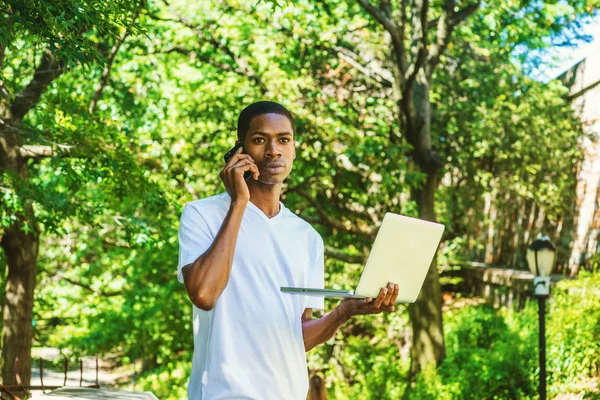 Multi Tasks.Young African American College Student Studying in New York, with short hair, wearing white T shirt, working on computer, talking on cell phone, walking under trees on campus in same time