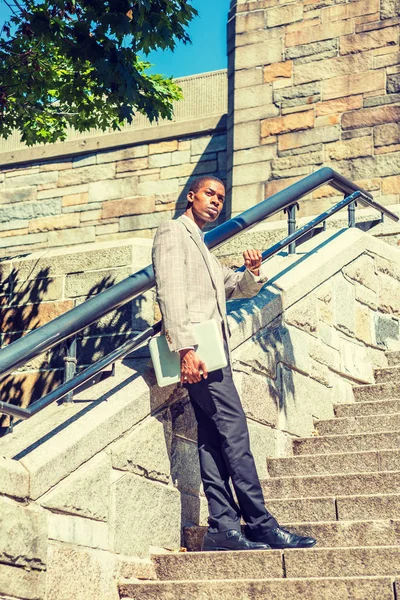 Time is Money. Young African American Businessman wearing patterned blazer, black pants, holding laptop computer, standing on rock stairs, raising arm with wristwatch, checking time, waiting, looking