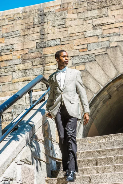 Young Happy African American Businessman traveling in New York City, wearing patterned blazer, white undershirt, black pants, leather shoes, walking down rock stairs by railing, looking around