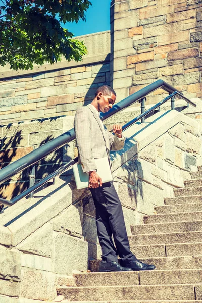 Time is Money. Young African American Businessman wearing patterned blazer, black pants, holding laptop computer, standing on rock stairs, raising arm with wristwatch, looking down, checking time