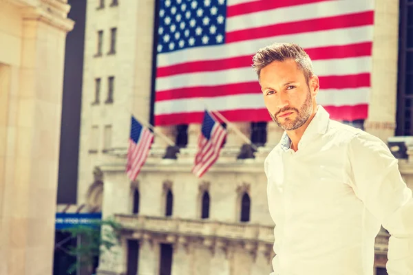 Young Sexy European Man with beard traveling in New York in summer, wearing long sleeve white shirt, standing outside old style office building with American flags under sun, looking forward