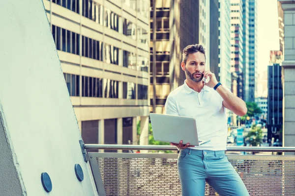 Young European Man working in New York City, with beard, little gray hair, wearing white Polo shirt, light blue pants, standing by railing on balcony, working on laptop computer, talking on cell phon