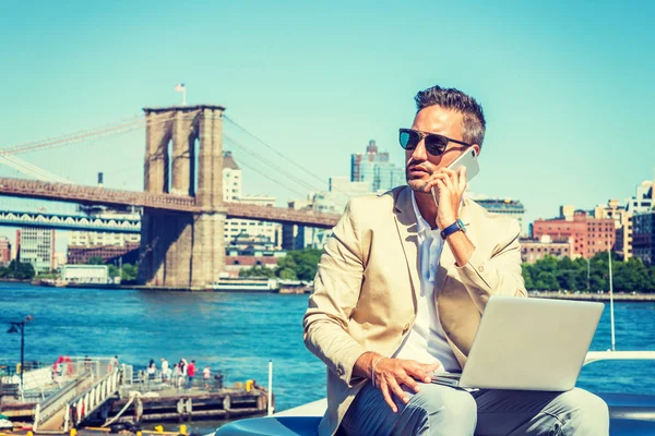 Avrupa genç işadamı seyahat, New York, küçük gri saç, sakal, çalışma bej blazer, güneş gözlüğü, Doğu Nehri, dizüstü bilgisayar üzerinde çalışan cep telefonuyla konuşurken başına oturmuş takıyor.